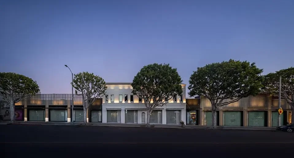 A low-rise retail block at dusk, continuous storefronts set behind mature street trees