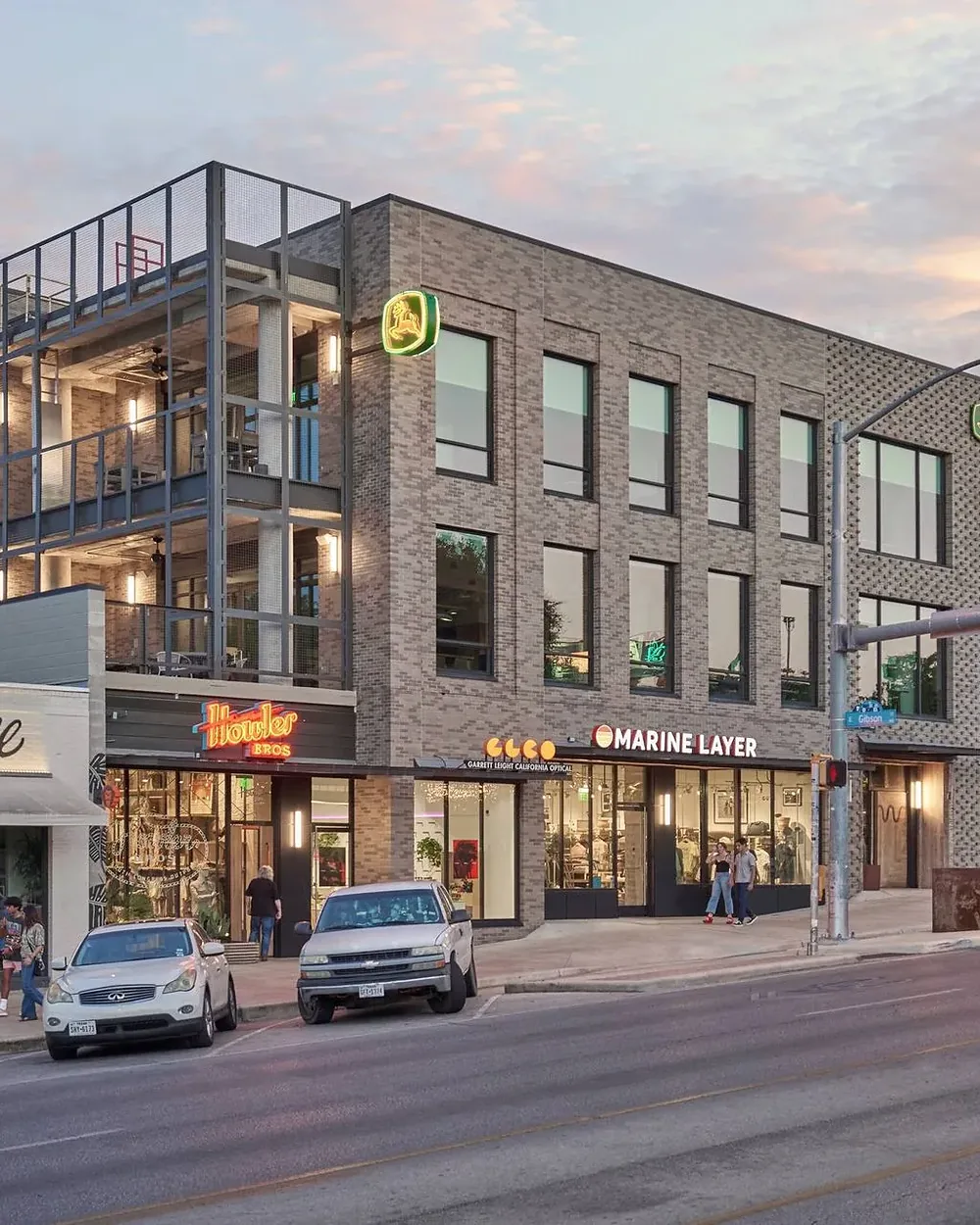 Dusk on a retail block, lit storefronts and people on the sidewalk