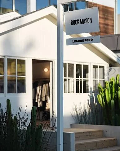 Buck Mason blade sign above a white cottage storefront and planted forecourt